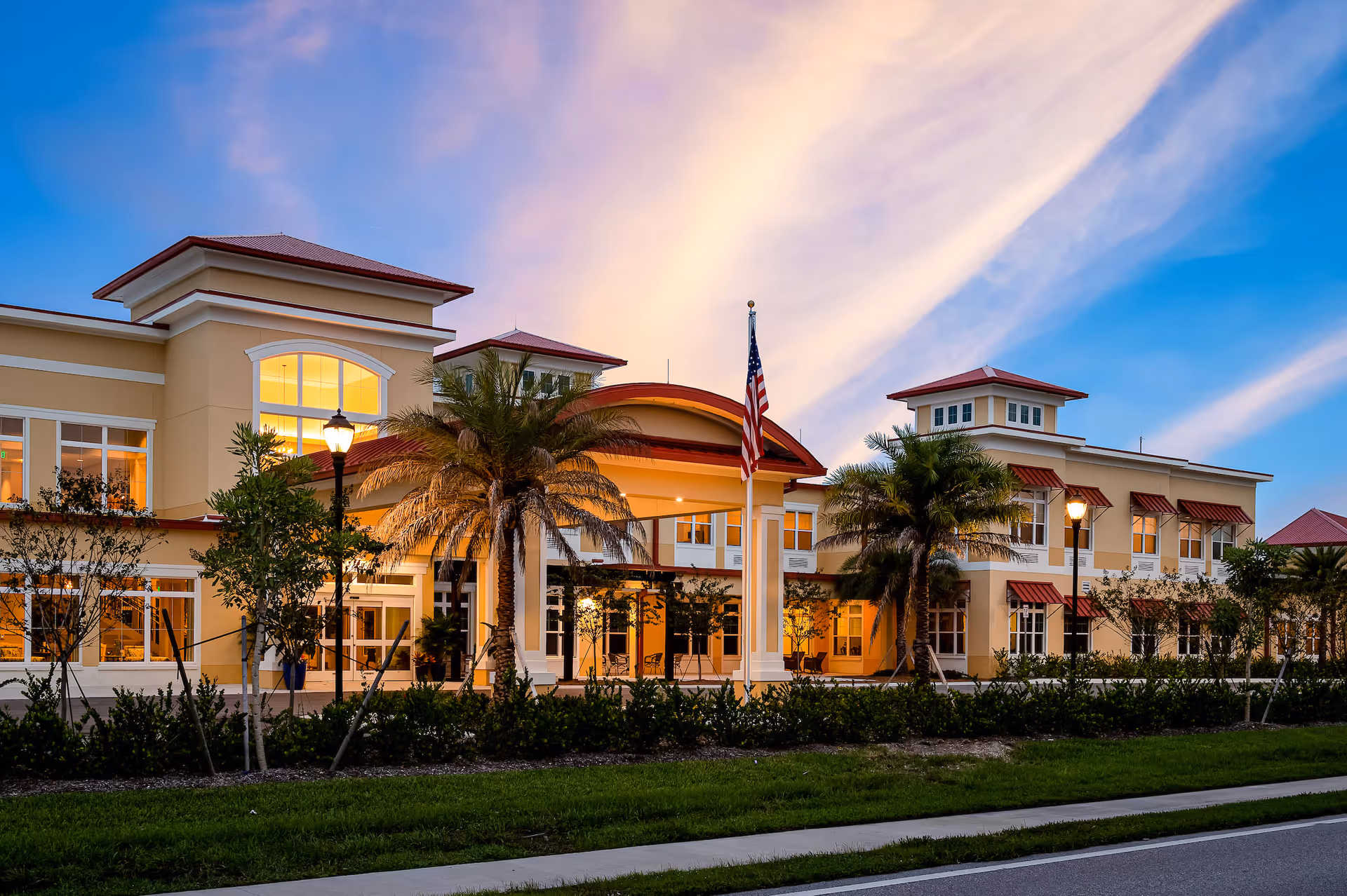 Exterior view of The Gallery at Cape Coral senior living facility at dusk, showing a large, well-lit building with palm trees, an American flag, and a clear sky with streaks of clouds.