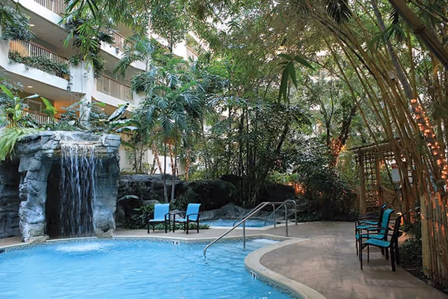 Indoor pool area with a waterfall feature, surrounded by lush tropical plants and trees. Several blue chairs are placed along the poolside and a pathway runs alongside the pool. The setting appears to be inside a building with balconies overlooking the pool area.
