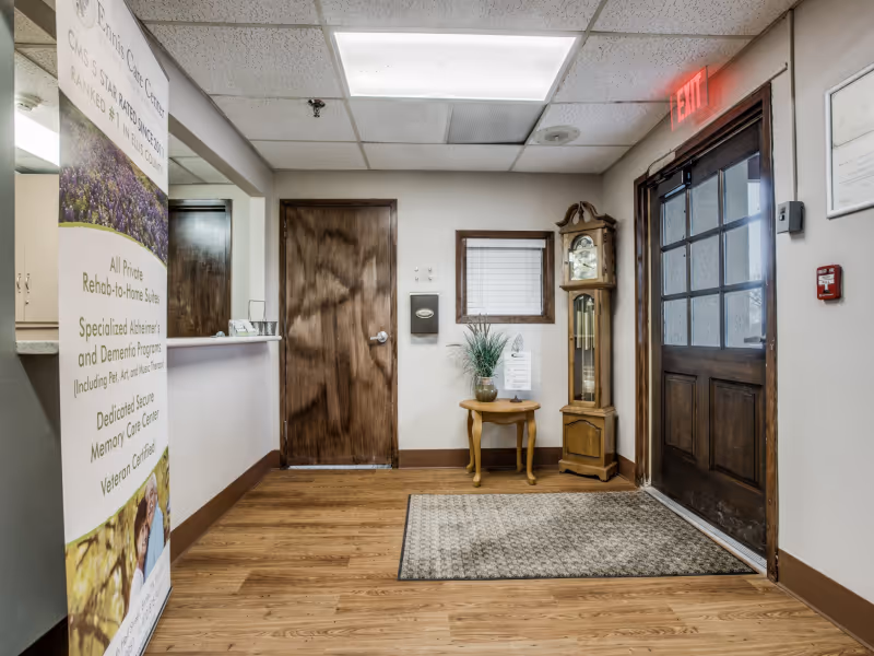 Interior view of the entrance area at Ennis Care Center featuring a wooden door with glass panels, an exit sign above, a grandfather clock, a small wooden table with a plant and a sign, a wooden door to another room, and a banner with information about the facility on the left side.