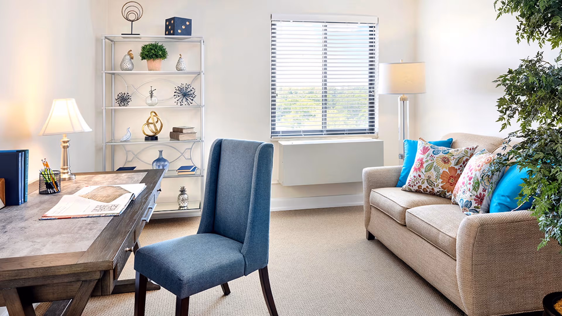 A bright and cozy living room with a beige sofa adorned with colorful floral and blue pillows, a blue upholstered chair, a wooden desk with papers and pencils, a metal shelving unit with decorative items, a window with blinds, and two lamps providing warm lighting.