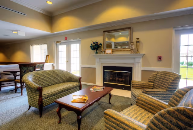 Cozy senior living lounge with upholstered sofas and armchairs arranged around a coffee table in front of a fireplace and French doors.