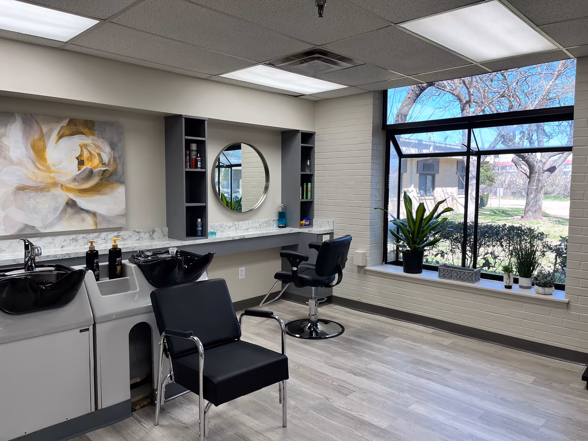 Interior view of a hair salon area with two black salon chairs, two black hair washing sinks, a round mirror mounted on the wall, shelves with hair products, a large window with plants on the windowsill, and a painting of a white and yellow flower on the wall.
