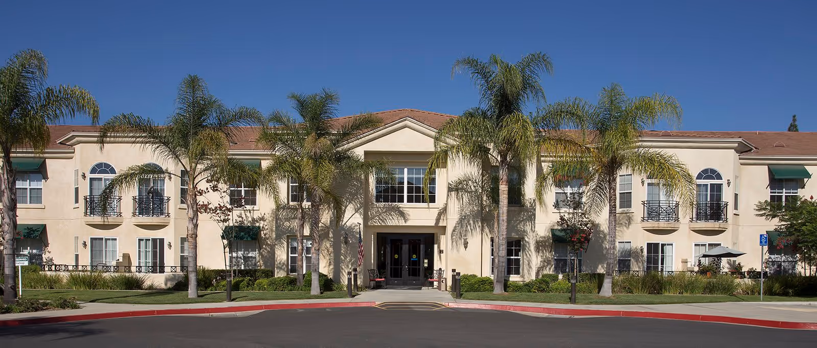 Front exterior view of a two-story retirement facility building with beige walls, red-tiled roof, multiple windows with green awnings, and several palm trees in front under a clear blue sky.