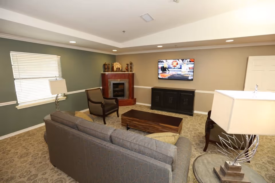 A cozy living room with a gray patterned sofa, two armchairs, a wooden coffee table, and a side table with a modern lamp. There is a fireplace in the corner with decorative items on the mantel, a flat-screen TV mounted on the wall above a black cabinet, and a window with blinds letting in natural light.