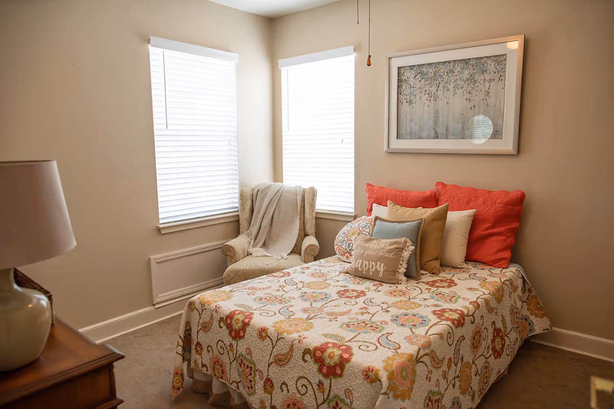 A cozy bedroom with a bed covered in a floral patterned quilt and multiple decorative pillows. There is a beige armchair with a throw blanket in the corner near two windows with white blinds. A framed artwork featuring trees hangs on the wall above the bed. A wooden nightstand with a lamp is partially visible on the left side.