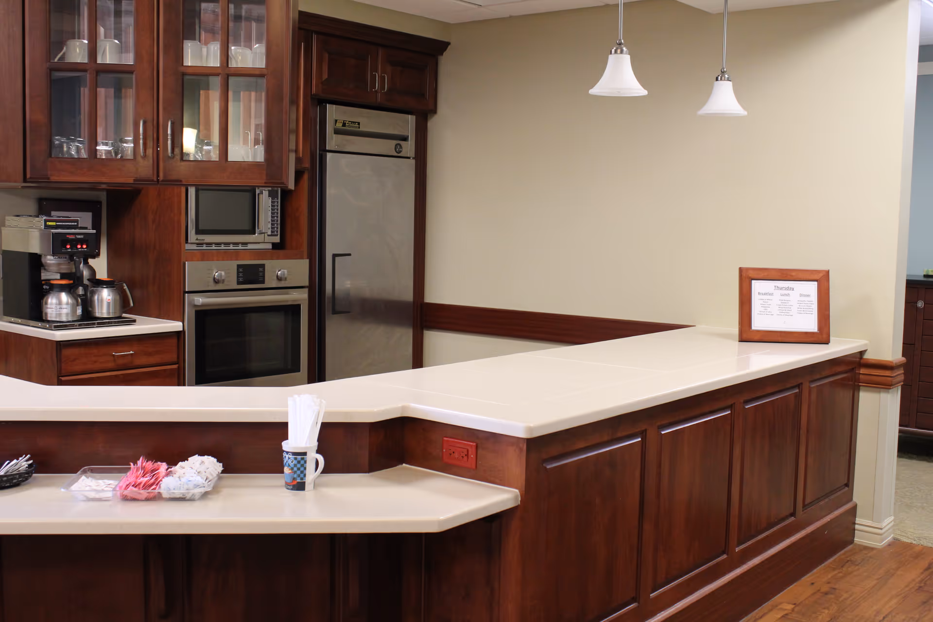 Warm-toned kitchenette with wooden cabinets, stainless appliances, a coffee station and a long white service counter.