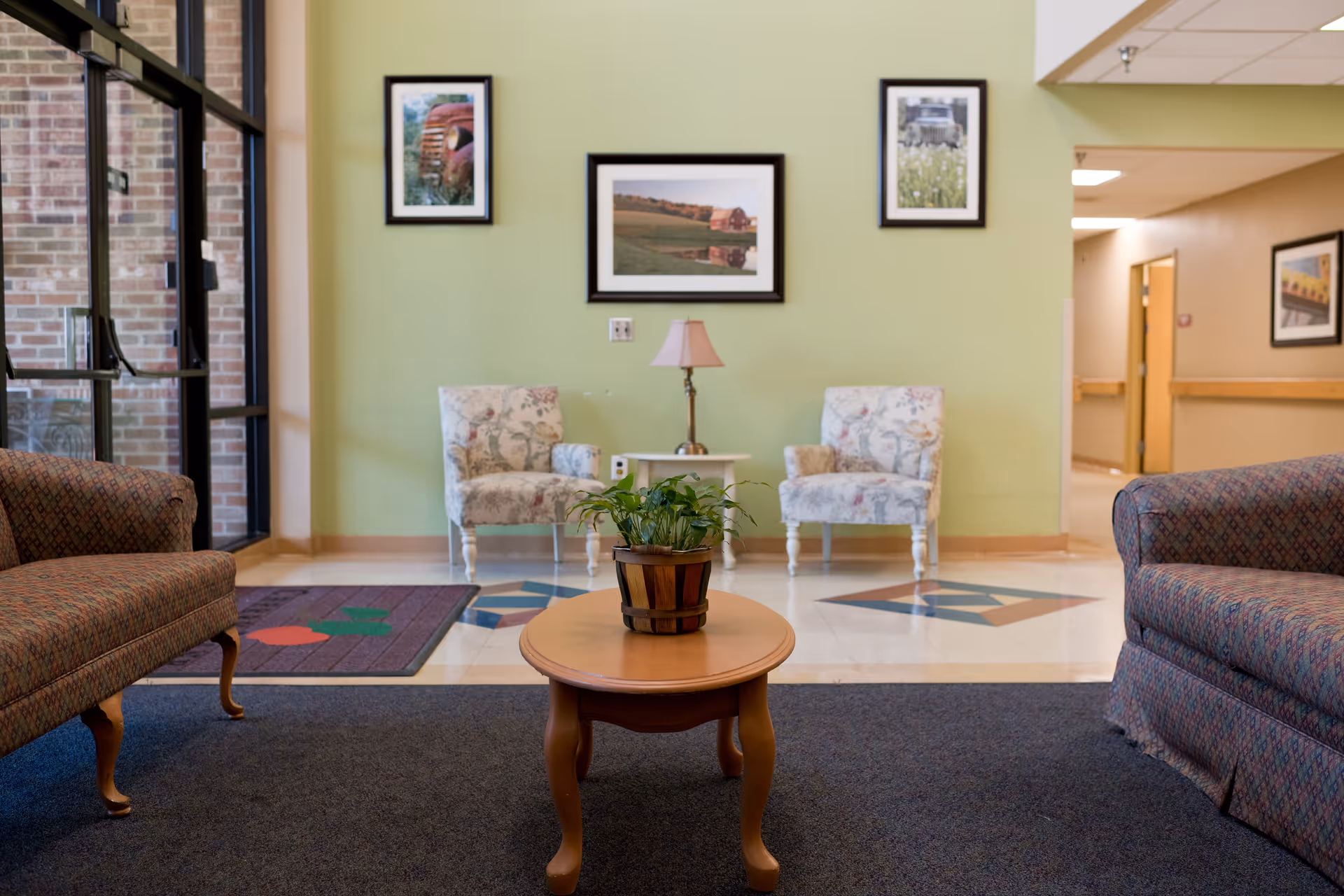 A cozy seating area in a senior living facility with two patterned armchairs and two sofas arranged around a wooden coffee table with a potted plant. The walls are light green with framed landscape pictures, and there is a small table with a lamp between the armchairs. The floor has a mix of carpet and tile with geometric designs.