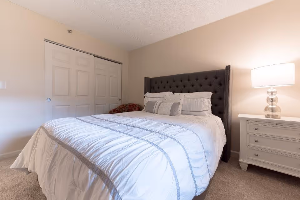 A bedroom with a large bed featuring a dark tufted headboard, white bedding with gray stripes, and several pillows. To the right of the bed is a white nightstand with a glass lamp on top. In the background, there is a closed closet with sliding white doors and a red patterned chair partially visible.