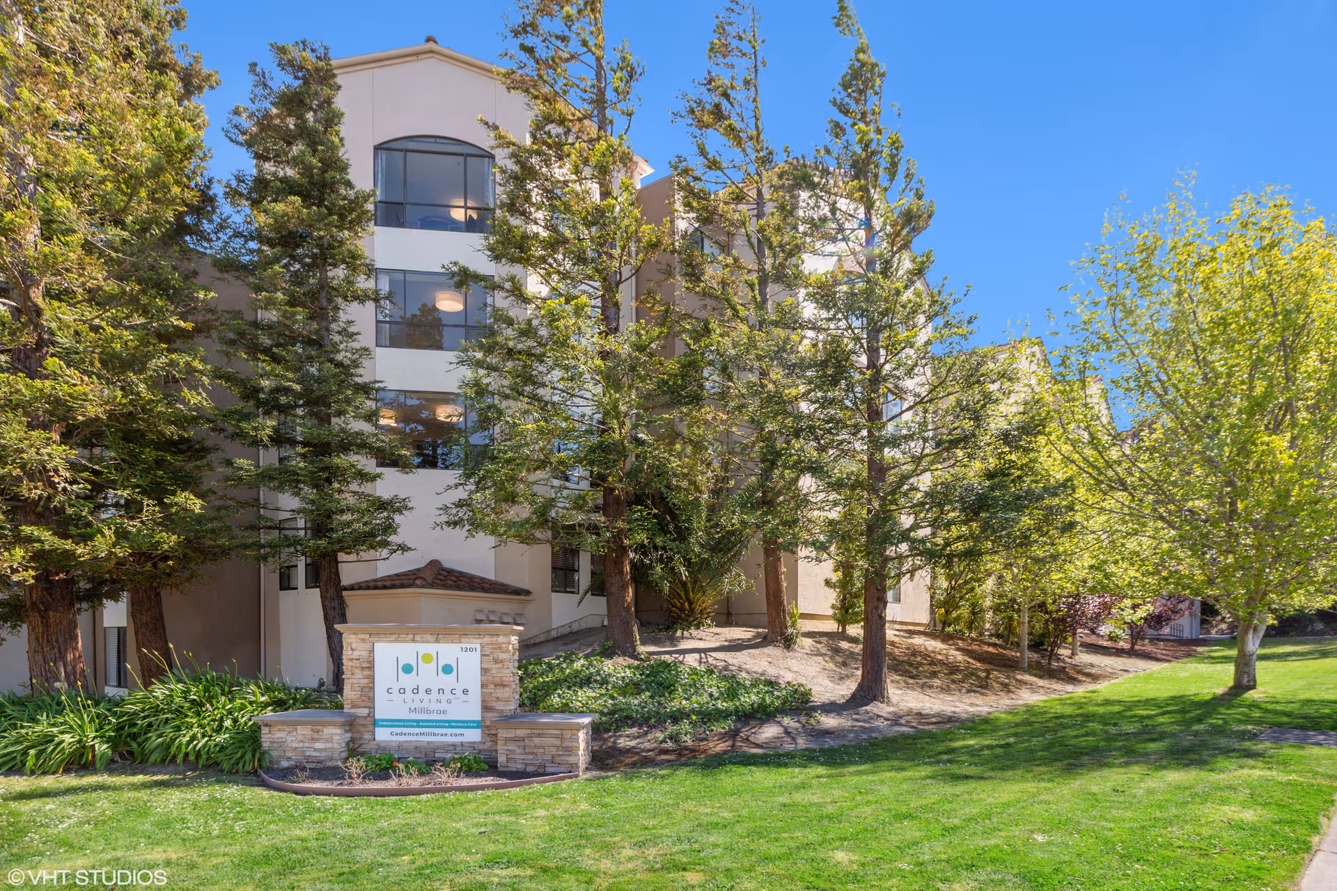 Exterior view of a multi-story senior living facility building surrounded by tall trees and green grass under a clear blue sky. A stone sign in front reads 'Cadence Living Millbrae' with the website cadenceMillbrae.com.