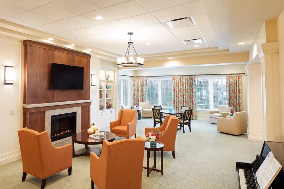 Sunlit communal lounge with a fireplace and wall-mounted TV, four orange armchairs around a coffee table, a dining table by large windows with floral curtains, and a piano.