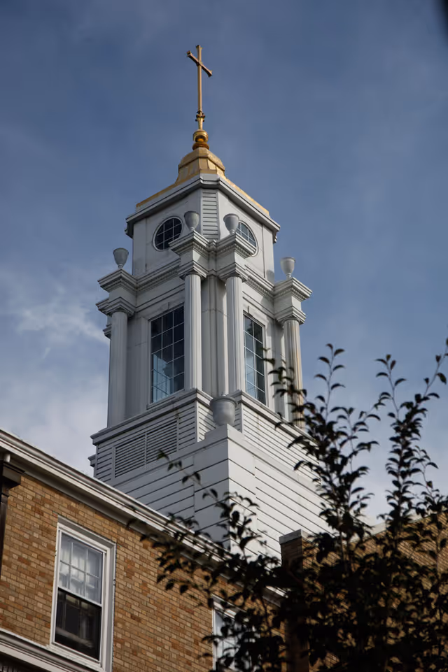 A white cupola topped with a gold cross rises above a brick building against a blue sky.