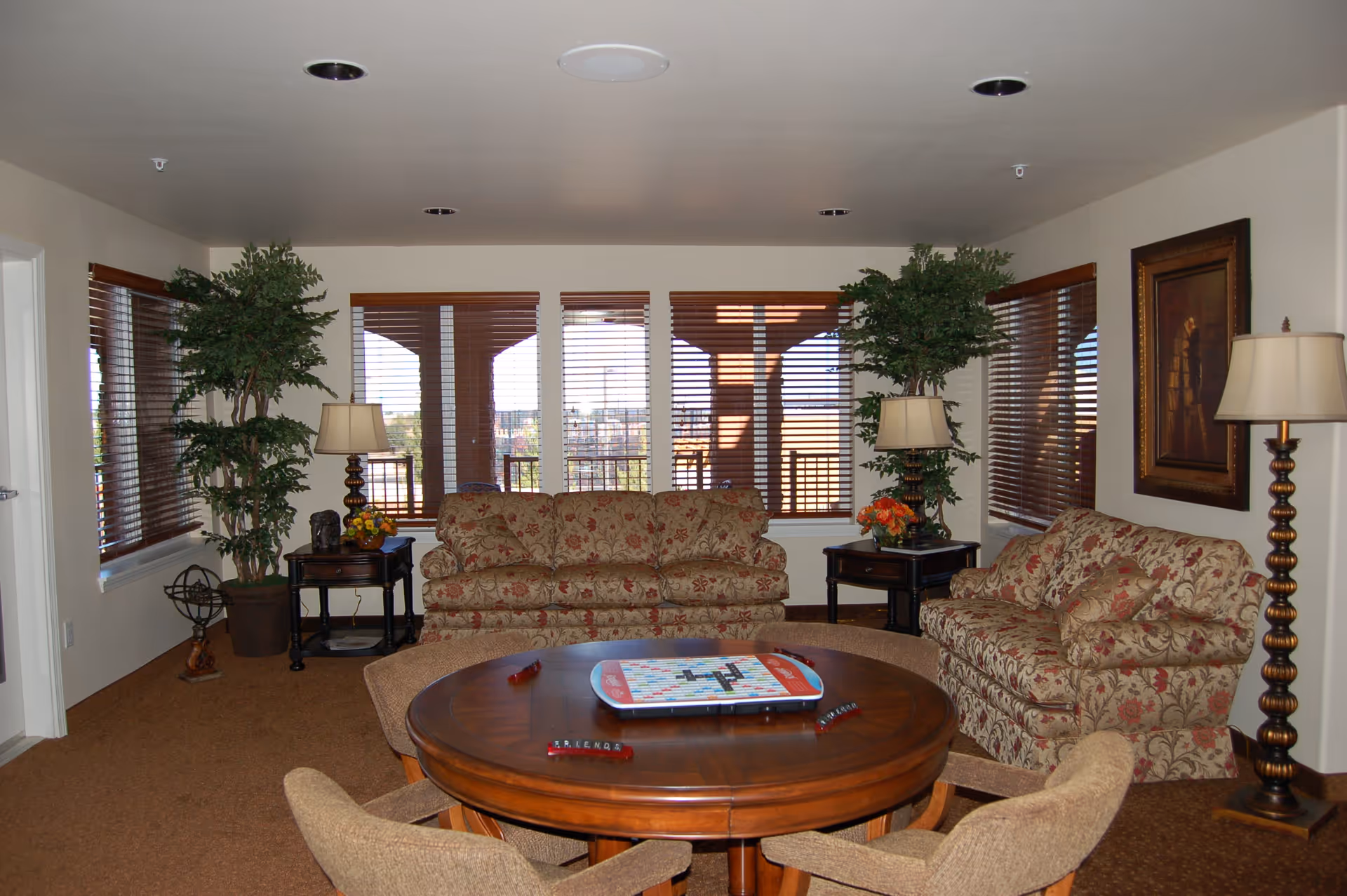 A cozy living room with two floral patterned sofas, a round wooden table with a Scrabble game set up, four beige upholstered chairs around the table, two tall potted plants, two side tables with lamps and flower arrangements, a floor lamp, and large windows with wooden blinds letting in natural light.