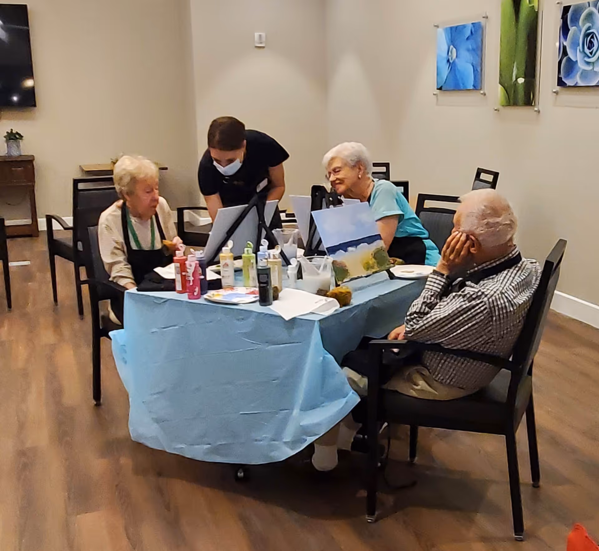 Three elderly individuals seated around a table covered with a blue tablecloth, engaging in a painting activity with canvases and paint bottles, assisted by a caregiver wearing a mask in a well-lit room with wooden flooring and floral wall art.