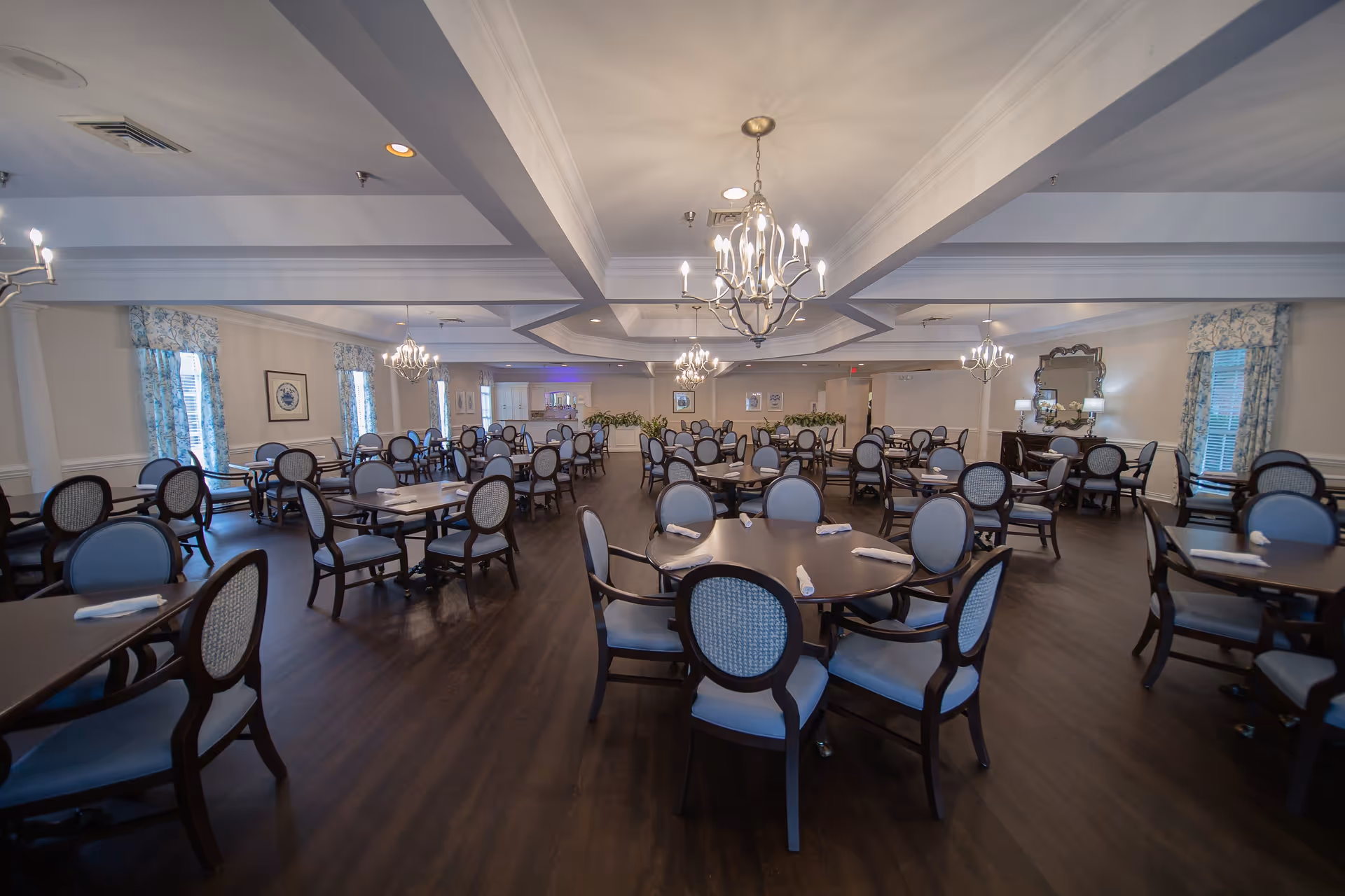 A spacious dining room with multiple round and rectangular tables surrounded by blue cushioned chairs. The room features chandeliers hanging from the ceiling, large windows with floral curtains, and a dark wooden floor. There is a sideboard with lamps and a mirror on the right wall.