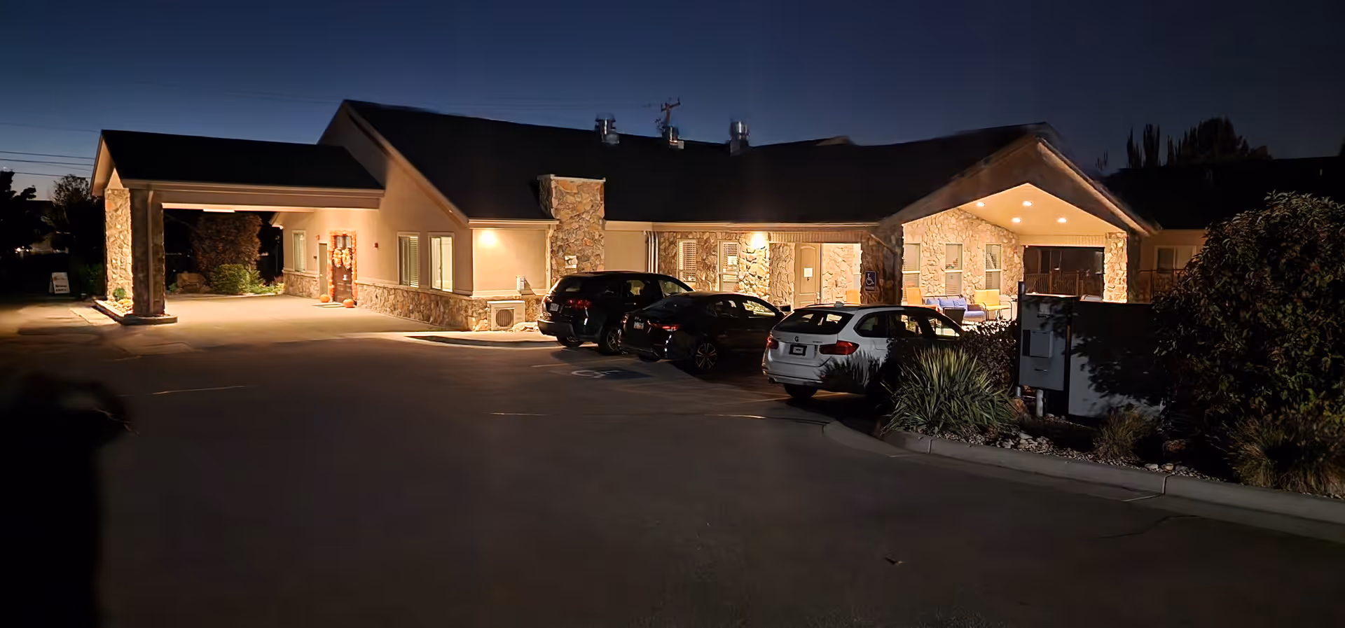 Nighttime view of the well-lit front entrance of a single-story senior living facility with several parked cars.
