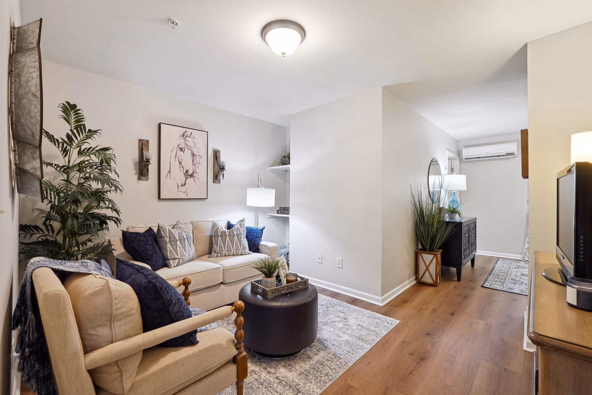 A cozy living room with a beige sofa adorned with patterned and solid blue pillows, a beige armchair with a blue throw blanket, a round dark ottoman serving as a coffee table with a tray and small plant, a large potted plant in the corner, wall art featuring a horse, and soft lighting from wall sconces and a table lamp. The room has light-colored walls and wood flooring extending into a hallway with additional furniture and decor.