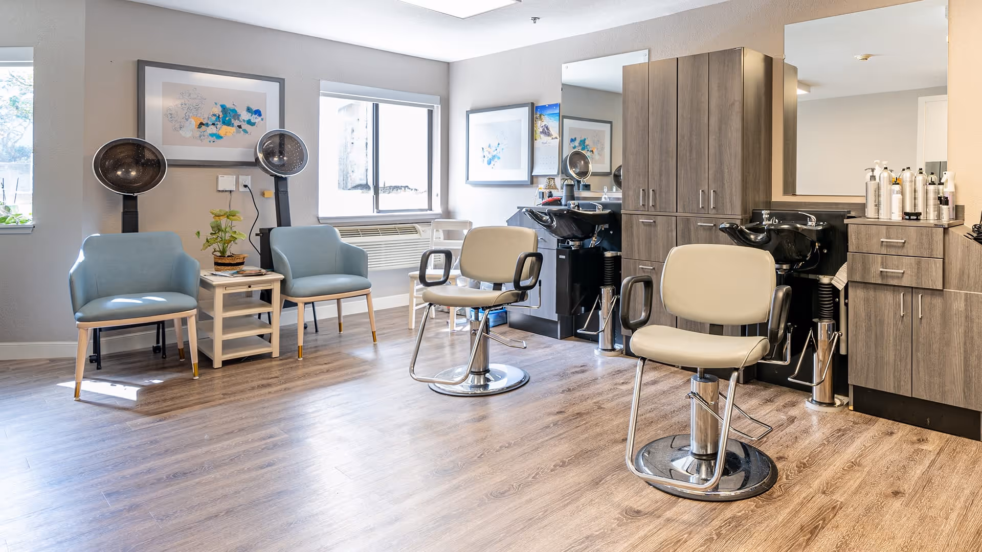 A bright and clean hair salon area inside a senior living facility with two salon chairs in front of wash basins and mirrors, two blue waiting chairs with a small table between them, and hair drying stations near the window. The room has wood flooring and light-colored walls with framed artwork.