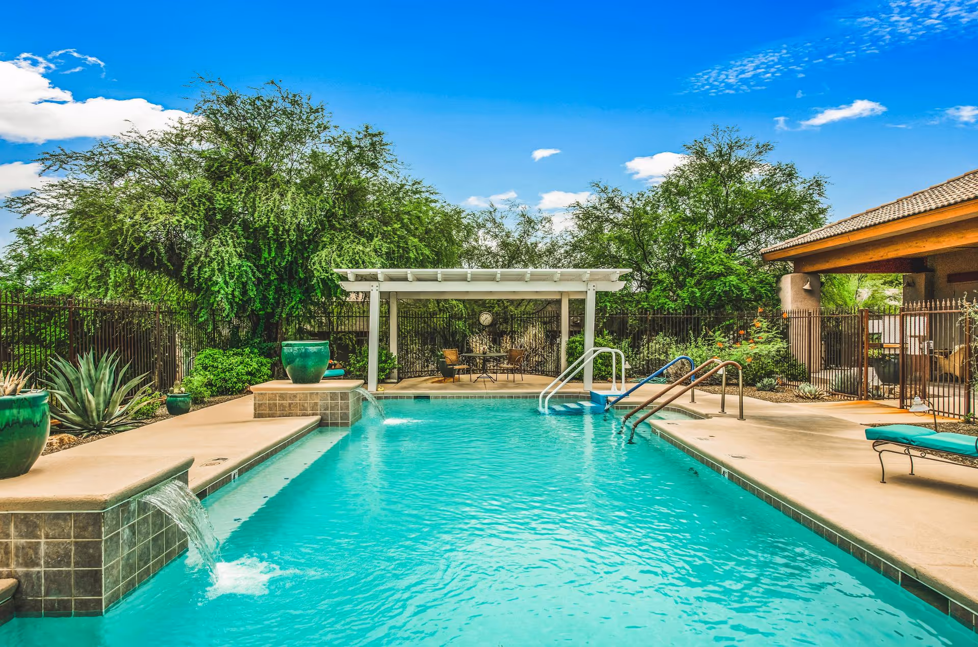 Outdoor swimming pool with clear blue water, surrounded by a concrete deck with lounge chairs and large green potted plants. There is a shaded seating area with chairs and a table at the far end of the pool, with trees and a bright blue sky with some clouds in the background.