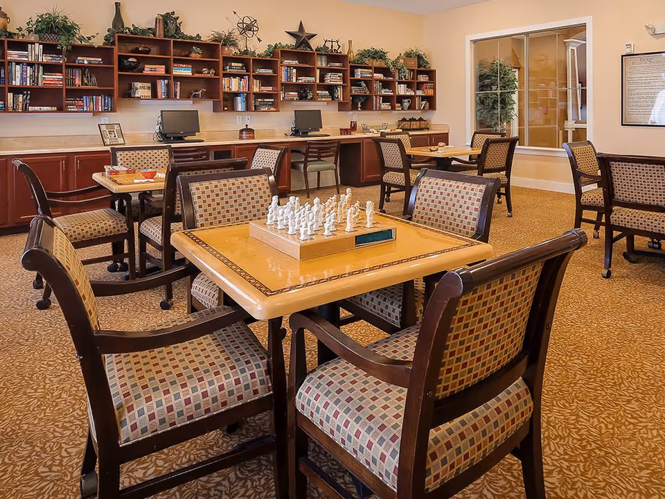 Community activity/dining room with tables and patterned chairs, a chess set on the nearest table, and bookshelves with computers along the back wall.