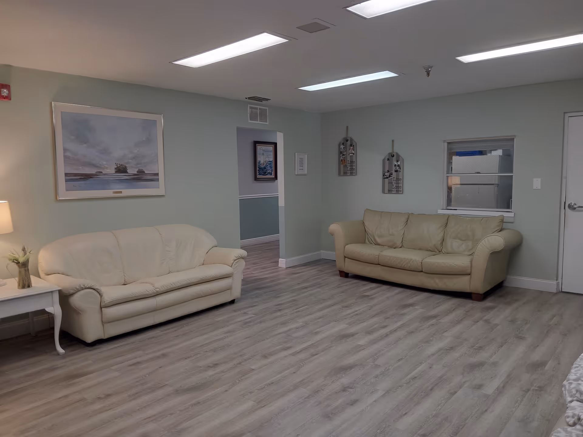 A living room area with two beige leather sofas facing each other, light wood flooring, a side table with a lamp and a small plant, a large framed painting on the wall, and two decorative wall hangings. The walls are painted light green and there is a small window and a door on the right side.