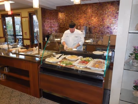 A chef wearing a white uniform and black hat prepares food behind a glass food display counter in a dining area. Various dishes are arranged on the counter, and the background features a wall with a mosaic tile design. The room has warm lighting and wooden furniture.
