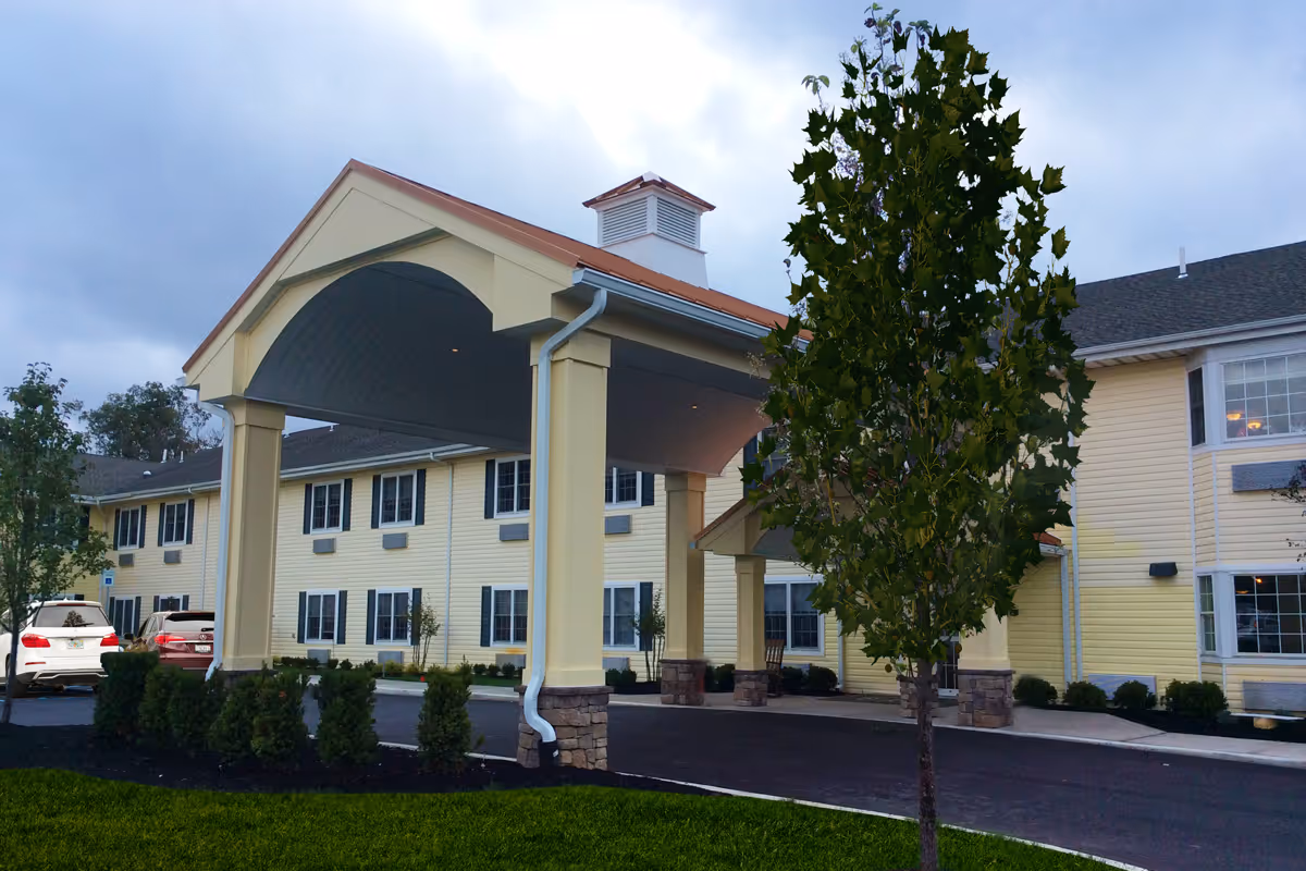 Exterior view of a two-story senior living facility building with a covered entrance driveway, several windows, and a tree in the foreground. There are parked cars visible near the entrance.