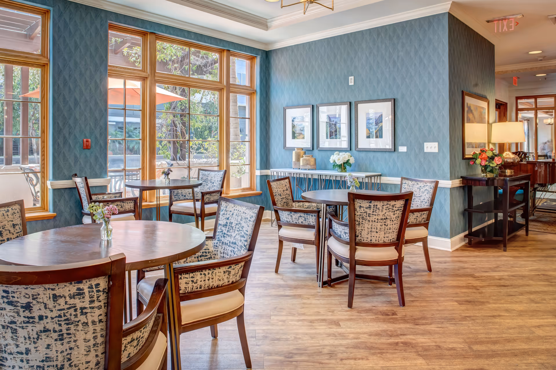 A bright dining area with several round wooden tables and cushioned chairs featuring patterned upholstery. Large windows with wooden frames allow natural light to fill the room, revealing an outdoor patio with umbrellas. The walls are painted blue with a textured pattern and decorated with framed artwork and floral arrangements. The floor is wooden, and there is a side table with a lamp and flowers near the hallway.