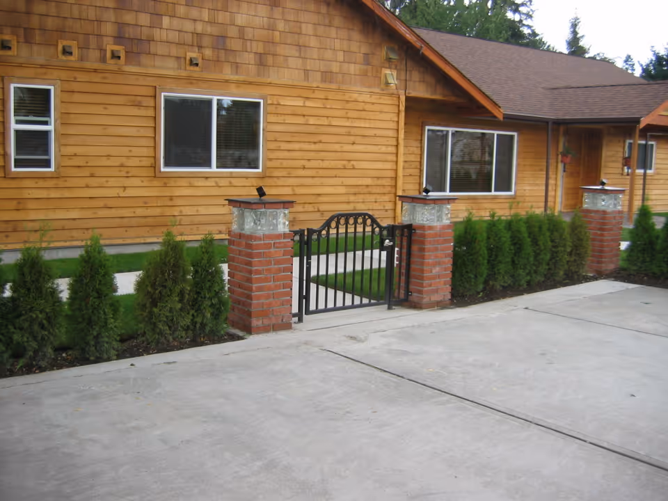 Exterior view of a wooden building with a brown shingled roof, featuring two windows and a door. In front of the building is a small black metal gate supported by two brick pillars with glass block tops, flanked by green shrubs and a concrete driveway.