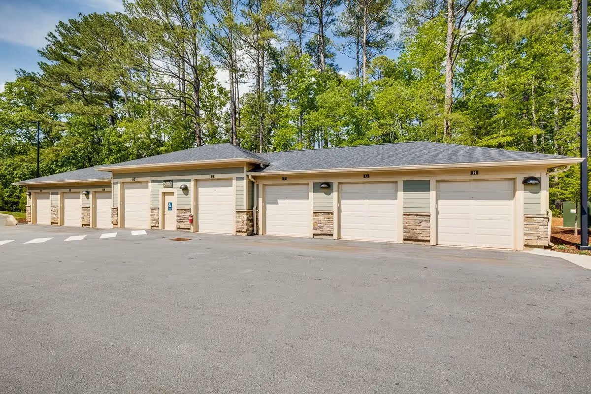Row of attached garage units with multiple white roll-up doors in front of a wooded backdrop.