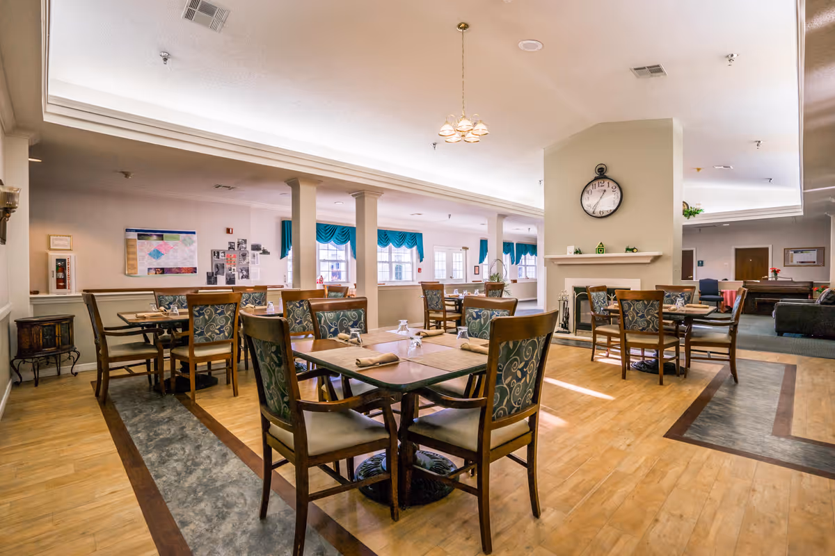 A bright and spacious dining room with multiple wooden tables and chairs arranged neatly. The room features a large clock on a central wall above a fireplace, large windows with teal curtains allowing natural light, and a wooden floor with decorative inlays. The space appears clean and welcoming, suitable for communal dining.
