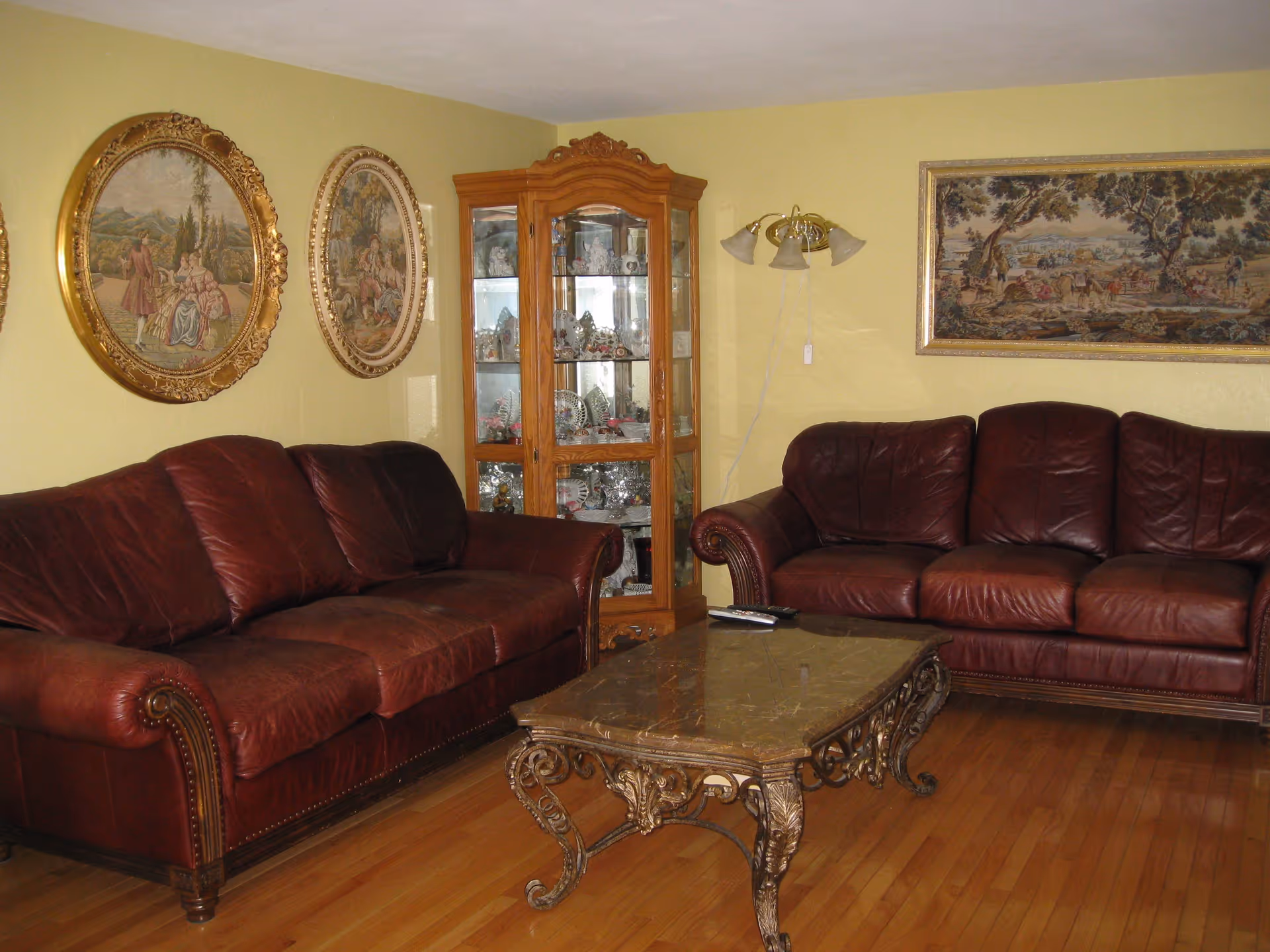 A living room with two burgundy leather sofas, an ornate coffee table, framed tapestry-style wall art, and a glass curio cabinet.