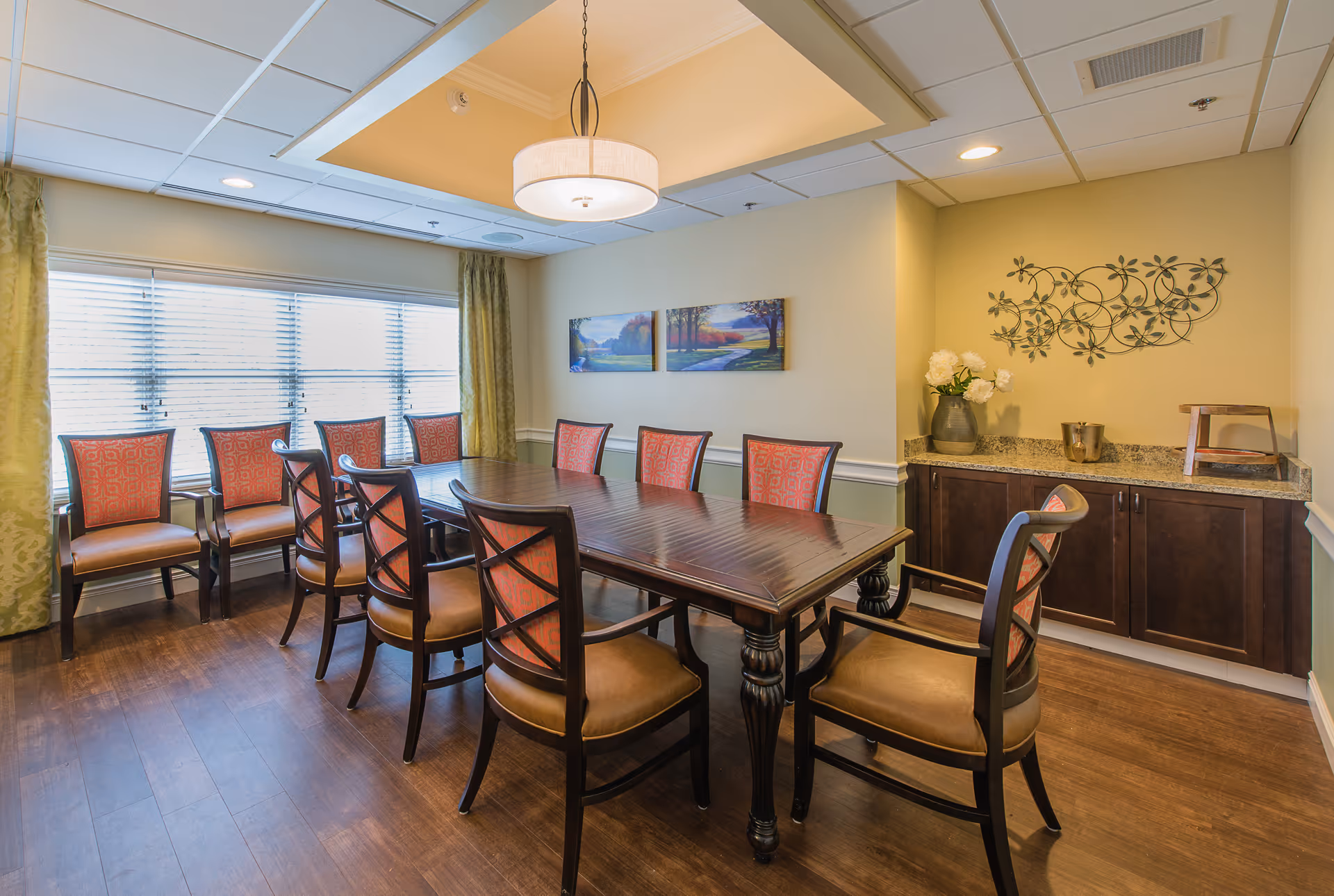 A well-lit dining room with a large rectangular wooden table surrounded by ten chairs with red patterned upholstery. The room has wooden flooring, a large window with blinds and green curtains, a ceiling light fixture, and wall decorations including a three-panel landscape painting and a metal floral wall art above a granite countertop with dark wooden cabinets.