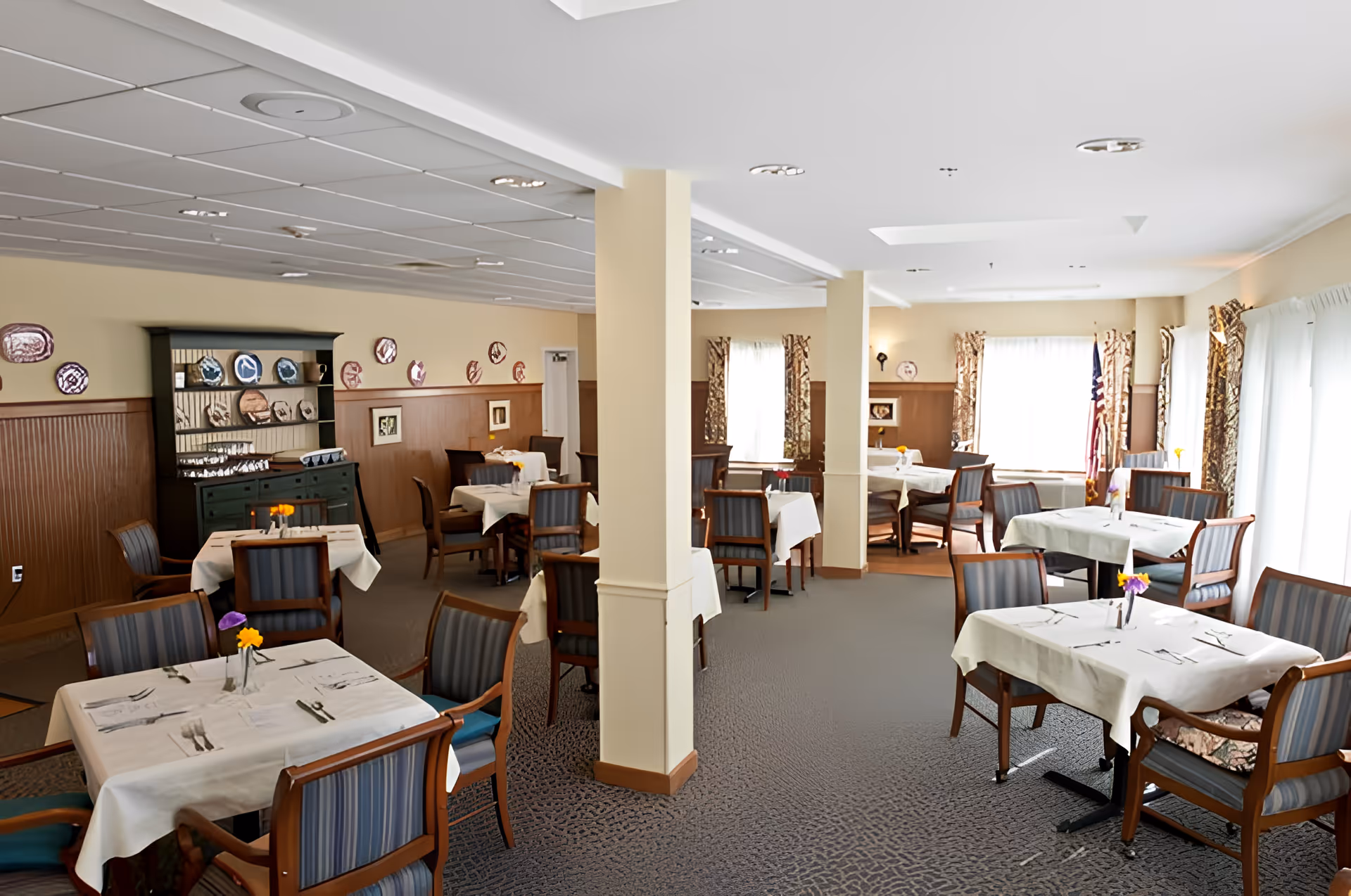 Bright dining room with several tables covered in white tablecloths and chairs arranged for meals.