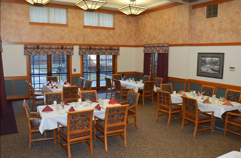 Dining room with multiple tables set with white linens and folded napkins, wooden chairs, large windows and patterned walls.