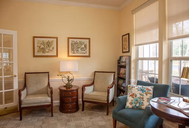 A cozy sitting area with two wooden armchairs with light upholstery, a round wooden side table with a lamp and a small plant, a blue upholstered armchair with a floral cushion, a round wooden table with magazines, and a rack with brochures near large windows with beige roller shades.