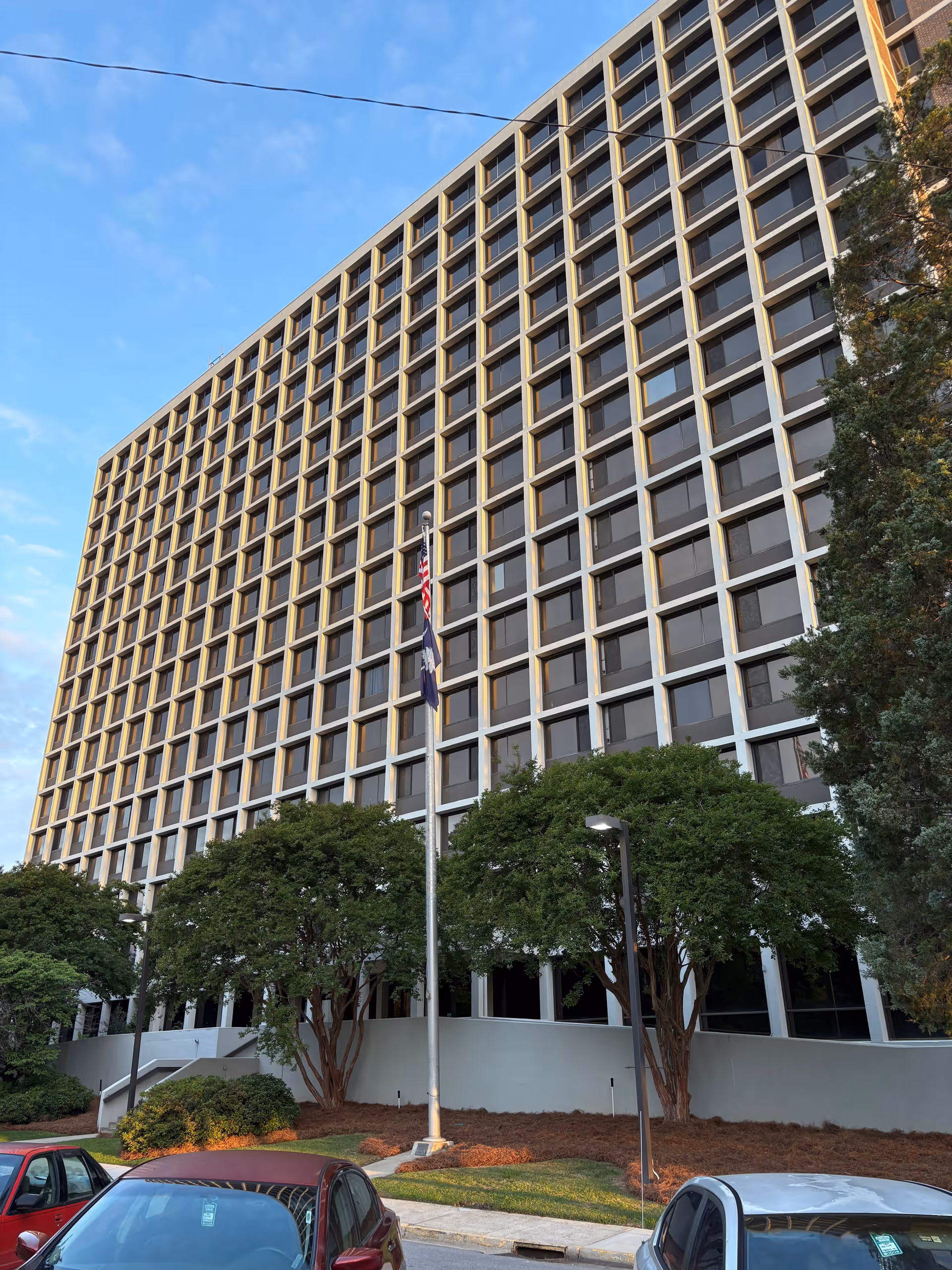 Exterior view of a multi-story building with a grid-like pattern of windows. In front of the building are several trees, a flagpole with two flags, and parked cars. The sky is partly cloudy with blue patches.