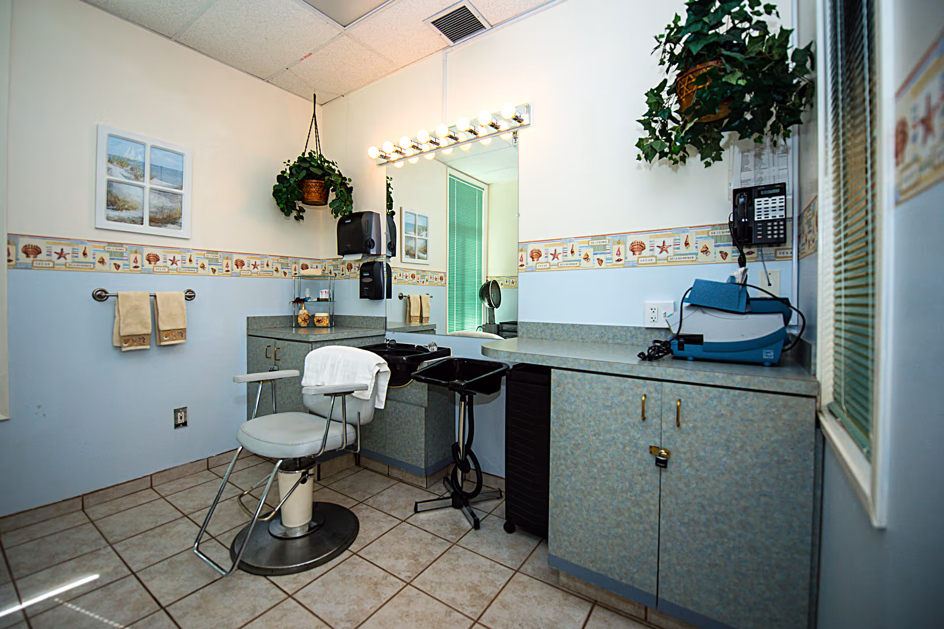 A small salon area inside a nursing center with a salon chair in front of a large mirror surrounded by vanity lights. There are two black sinks for hair washing, cabinets with a speckled blue-gray finish, a wall-mounted phone, and two hanging potted plants. The walls are decorated with a seashell and starfish border wallpaper and framed beach-themed pictures. The floor is tiled with light-colored tiles.
