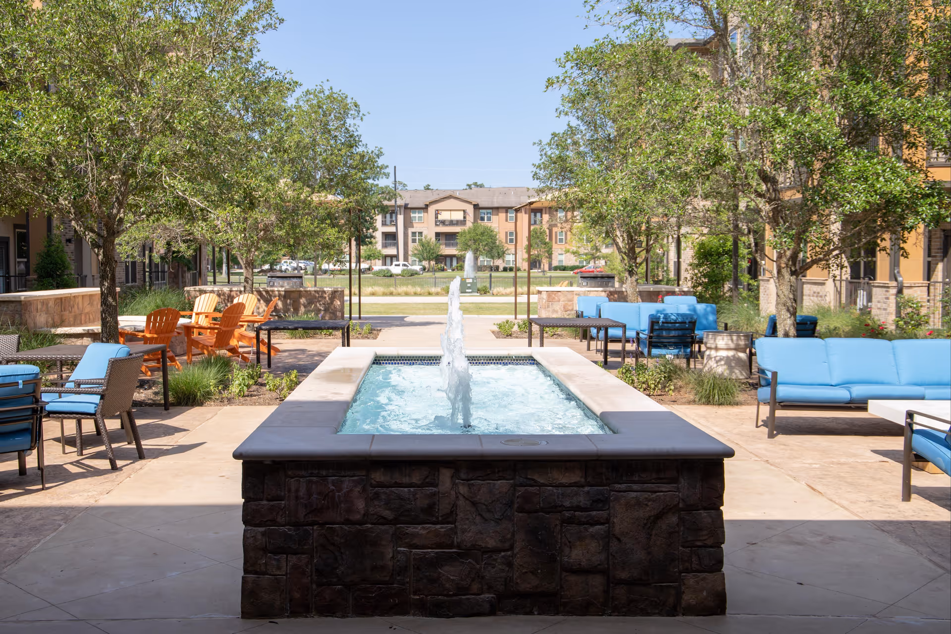 Outdoor courtyard area with a rectangular water fountain in the center, surrounded by trees, blue cushioned seating, and orange Adirondack chairs under a clear blue sky.