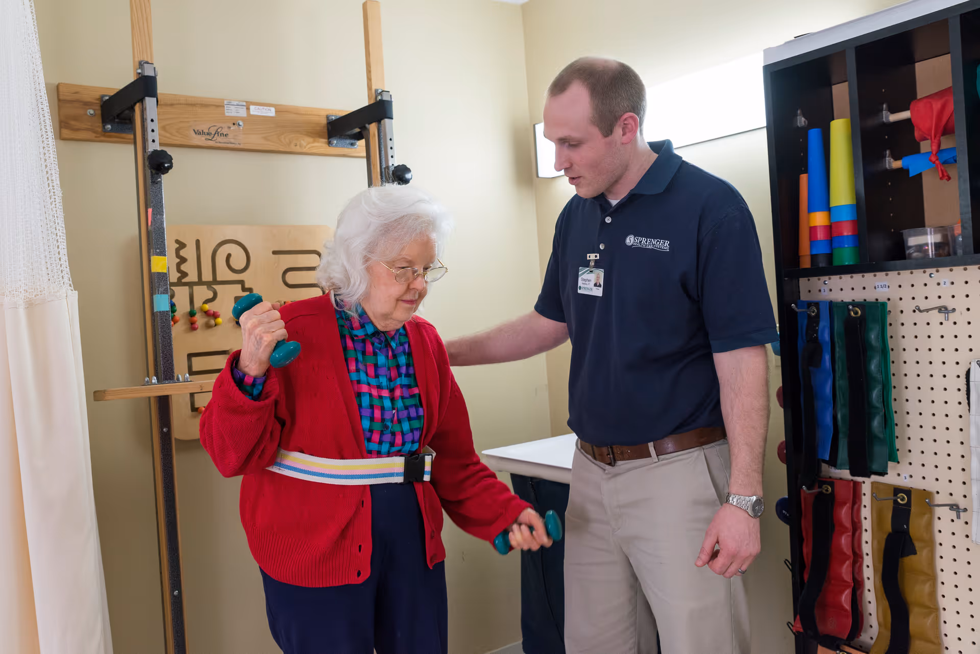 An elderly woman wearing a red cardigan and glasses is exercising with small dumbbells while a male healthcare worker in a navy blue Sprenger Healthcare Massillon polo shirt assists her in a therapy room with exercise equipment and therapy tools on the wall.