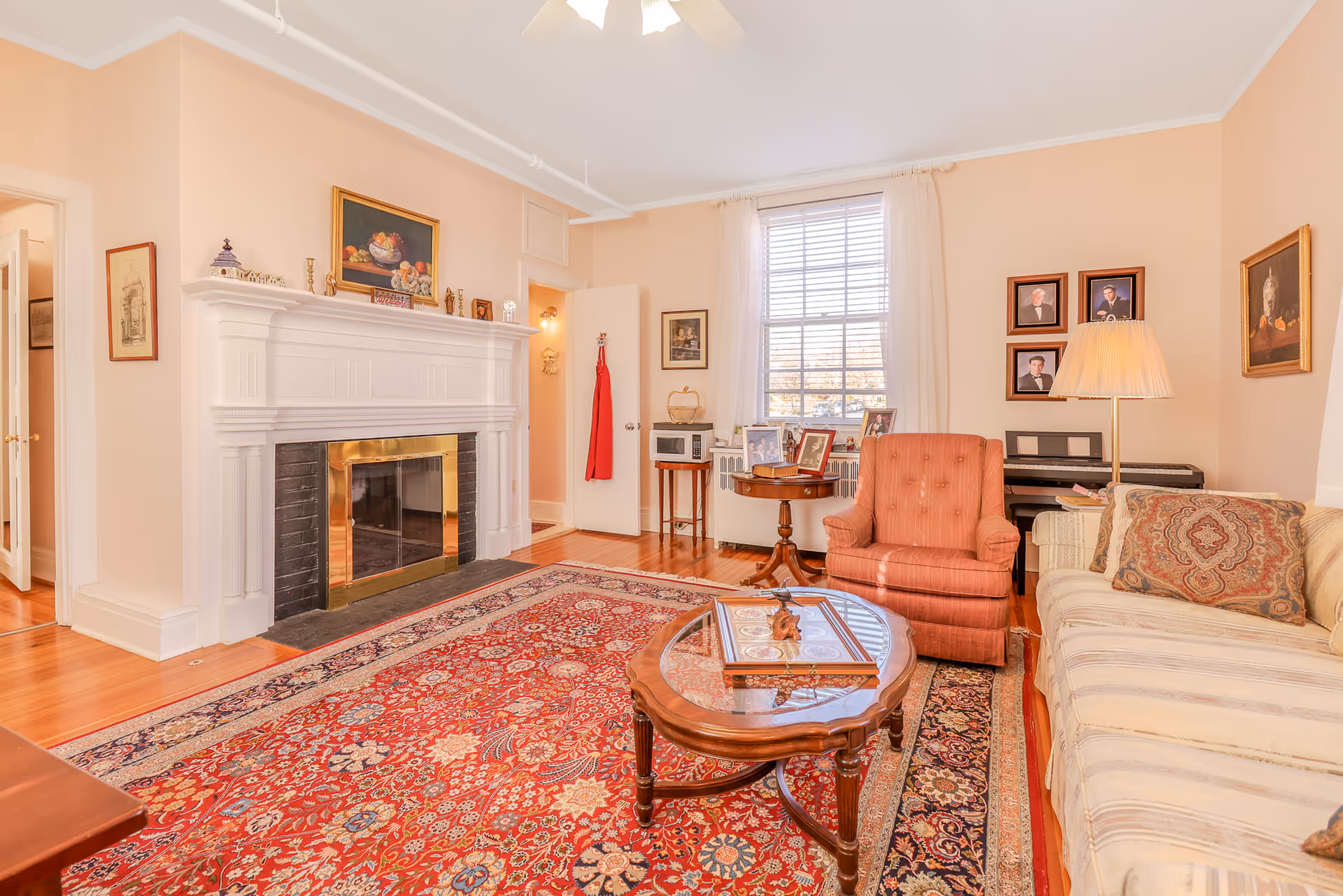 A cozy living room with a large red patterned rug, a wooden coffee table with glass top, a beige striped sofa with patterned cushions, and a coral upholstered armchair. There is a white fireplace with a brass screen, decorated with small items and a painting above it. A window with white curtains lets in natural light. The room also features a piano with framed photos on the wall above it, a standing lamp, and hardwood floors.