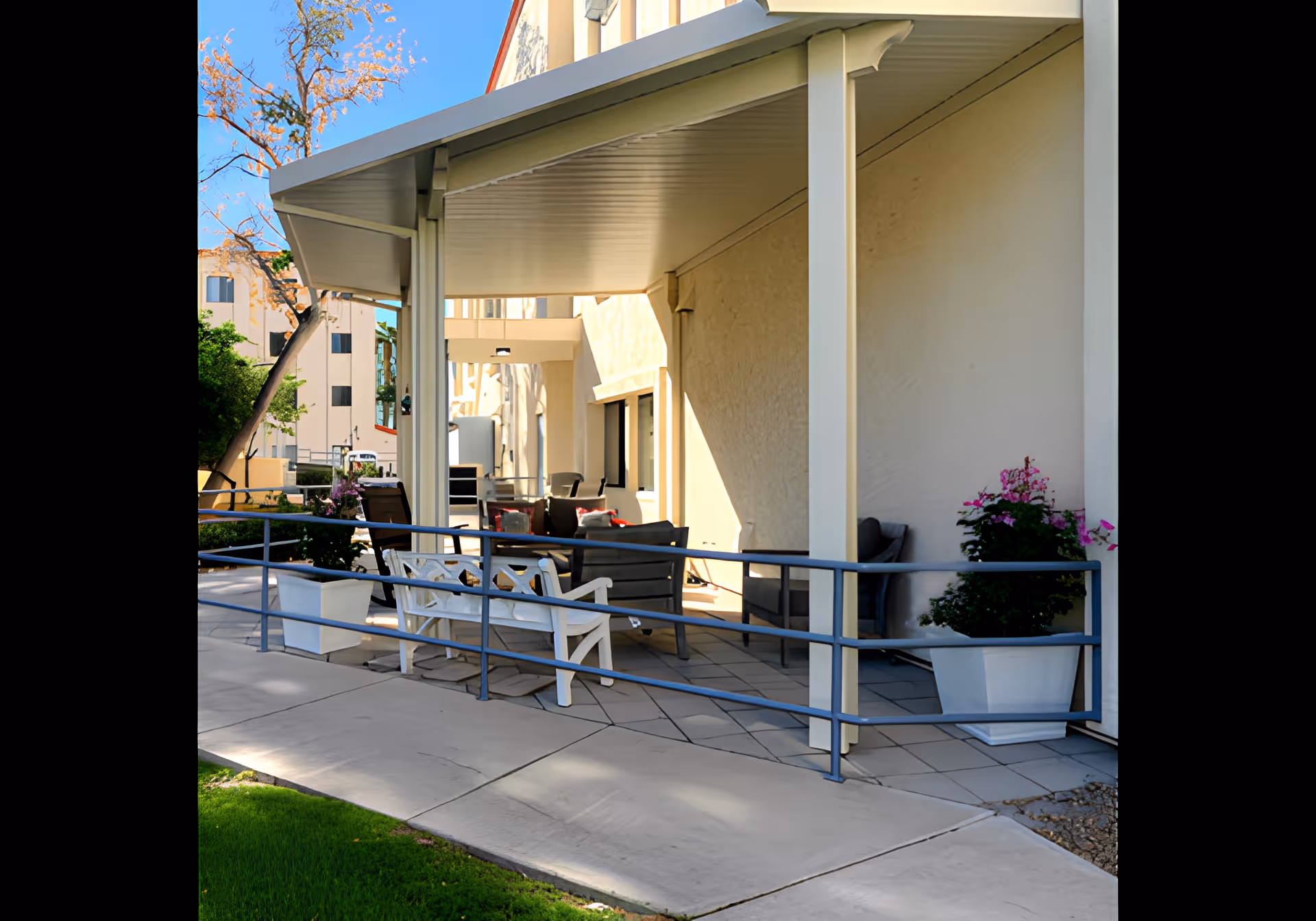 Covered outdoor patio area with seating including white benches and cushioned chairs, potted plants with pink flowers, and a metal railing. The patio is adjacent to a beige building with windows and is surrounded by greenery and other buildings in the background.