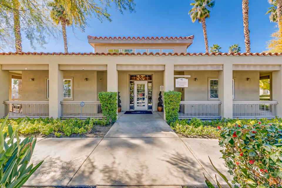 Front exterior view of a single-story building with a tiled roof and a covered porch area. The entrance has glass double doors flanked by potted plants and greenery on either side. There are palm trees and other plants around the building under a clear blue sky.