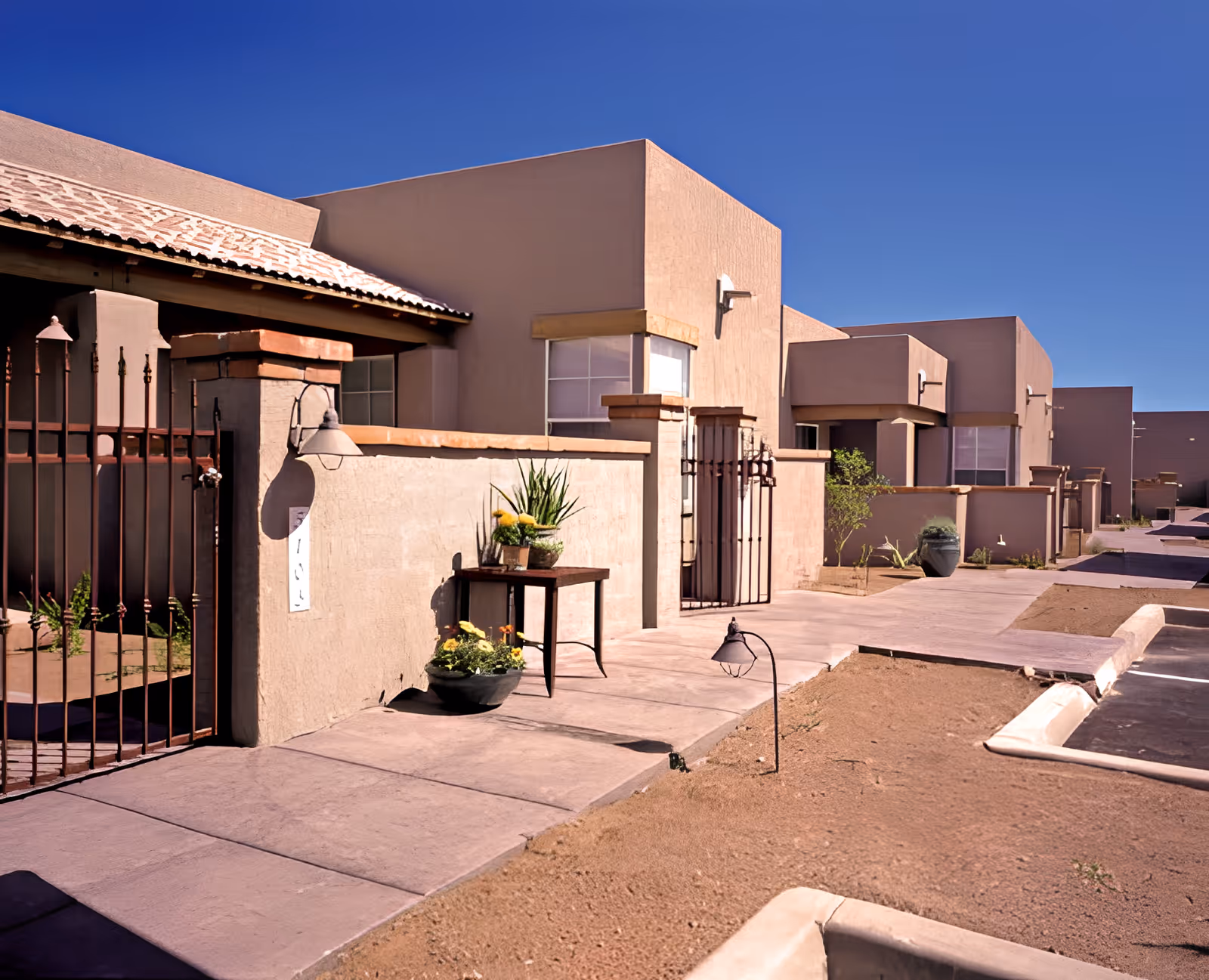 Row of stucco units with gated patios, potted plants, and a paved walkway under a clear blue sky.