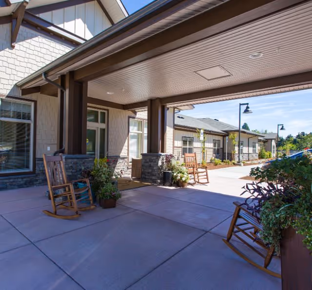 Covered outdoor patio area at a senior living facility with wooden rocking chairs and potted plants. The building exterior features stone accents and beige siding under a clear blue sky.