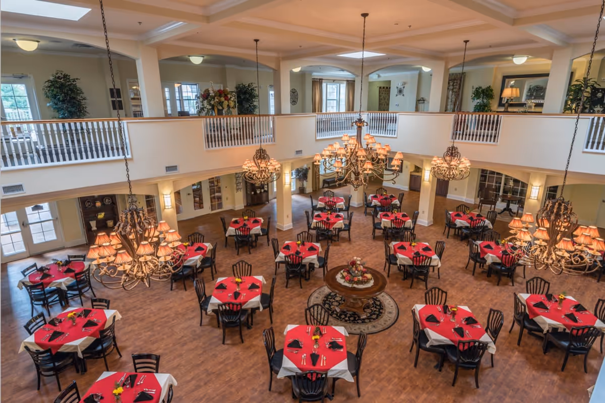 Spacious two-story dining room with multiple round tables set with red tablecloths, ornate chandeliers, and a central decorative table.