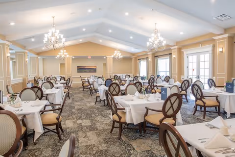 A spacious dining room with multiple tables covered in white tablecloths, each set with plates, napkins, and glassware. The room features elegant chandeliers hanging from a vaulted ceiling, beige walls with white trim, large windows allowing natural light, and patterned carpet flooring. Chairs with cushioned seats and backs surround each table.