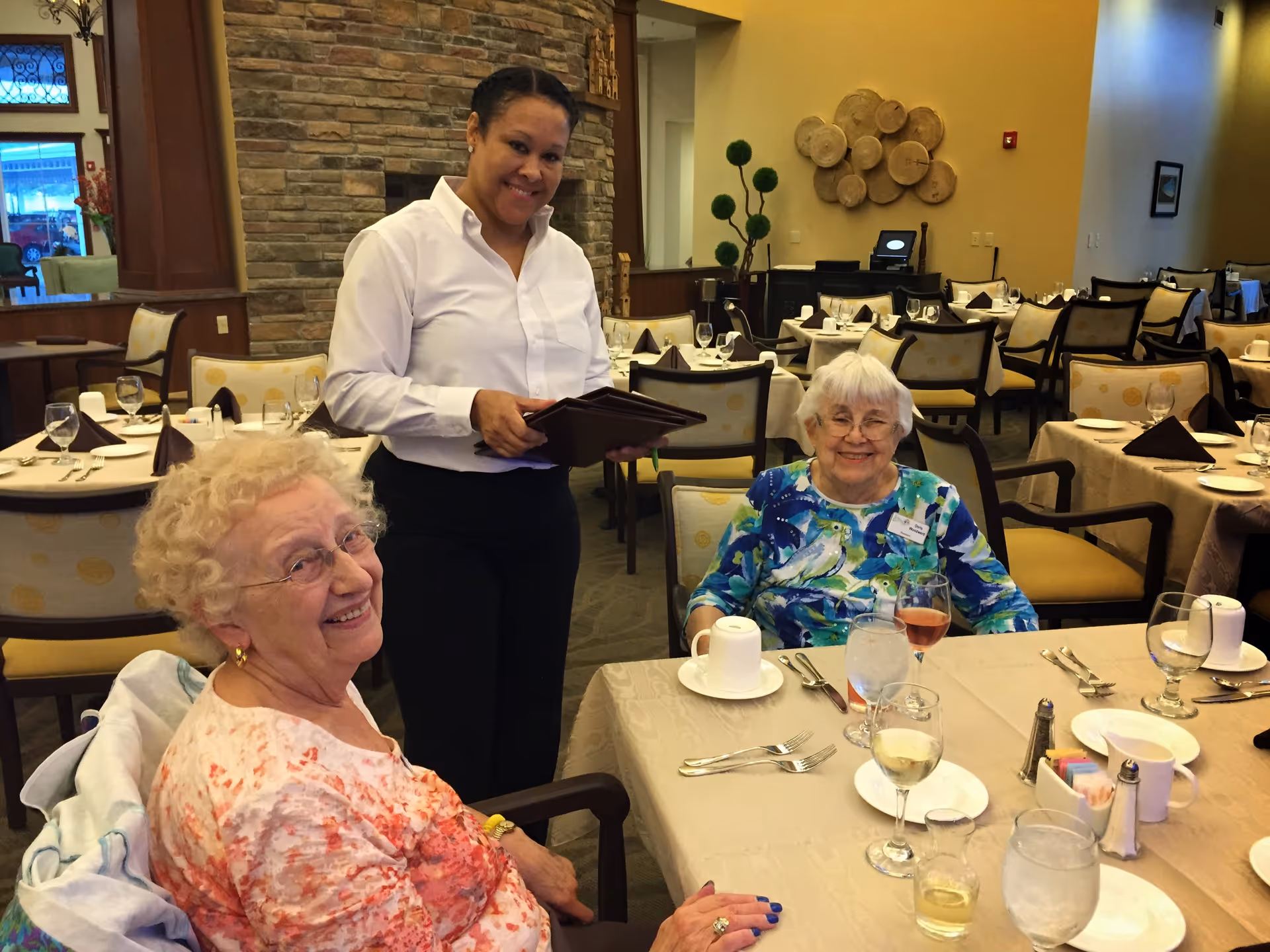 Two elderly women sitting at a dining table set with glasses, plates, and utensils in a senior living facility dining room. A smiling waitress in a white shirt and black pants stands beside them holding a menu. The room has multiple tables and chairs, a stone fireplace, and decorative wall art.