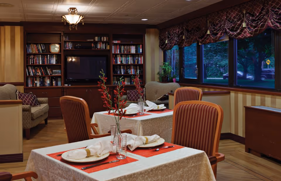 A cozy dining area with two tables set for a meal, each with white tablecloths, red placemats, plates, napkins, and glasses. Behind the tables, there is a living space with armchairs, a large bookshelf filled with books, and a television. Large windows on the right side show a view of trees and a parked car outside. The room has warm lighting and a comfortable, inviting atmosphere.