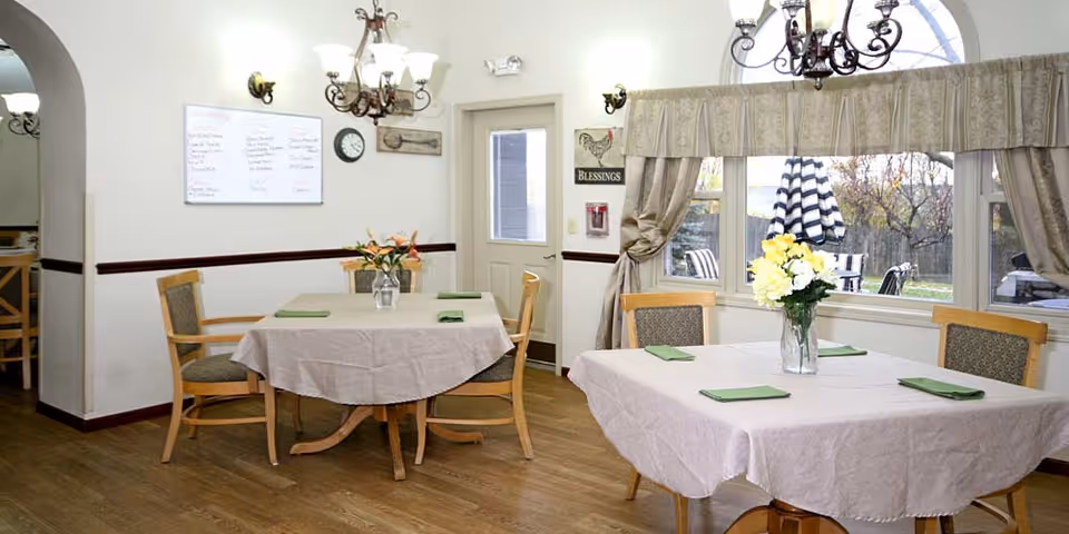 A bright dining room with two tables covered in beige tablecloths, each set with four green napkins and a vase of flowers. The room has wooden chairs, hardwood floors, a chandelier, and large windows with curtains showing an outdoor patio area. A whiteboard and a clock are mounted on the wall near a door.