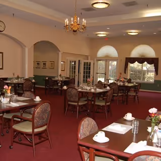 A dining room with multiple tables and chairs arranged neatly. Each table is set with white cups, glasses, napkins, and small flower vases. The room has red carpet flooring, green lower walls with beige upper walls, and arched doorways. There is a chandelier and ceiling lights providing illumination, and large windows with curtains letting in natural light.